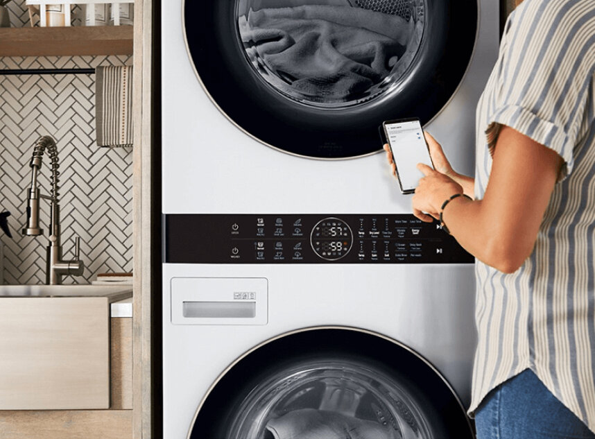 A person using the LG ThinQ® app on a smartphone in front of an LG Studio washer, emphasizing the reliability and 2-year warranty of LG appliances.