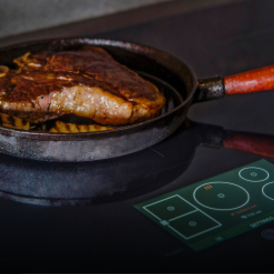 A steak searing in a hot cast iron pan on an induction cooktop with temperature controls visible. The word 'Induction' is displayed in the bottom corner.
